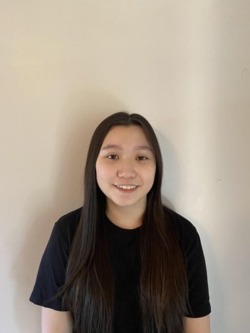 A young asian woman with long dark hair wears a black t-shirt and smiles in front of a beige wall.